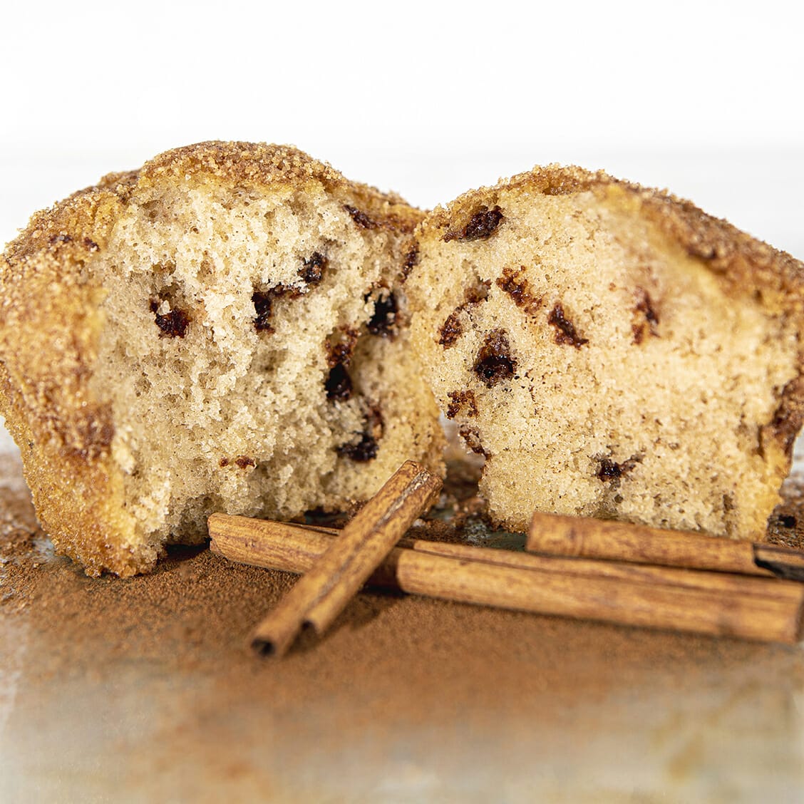 Cinnamon Chip gluten-free muffin broken open and surrounded by cinnamon sticks and cinnamon powder